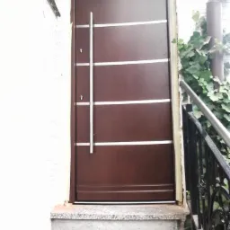 Ciemnobrązowe drzwi wejściowe z czterema poziomymi, srebrnymi paskami i długim, pionowym uchwytem, zamontowane na schodach z jasnego kamienia.