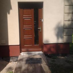 Zakład Stolarski Marcin Mateja - Częściowo otwarte, brązowe drzwi wejściowe z przeszkleniem i panelem, zamontowane w beżowej elewacji budynku z brązowym cokołem. Widoczne drewniane podesty.