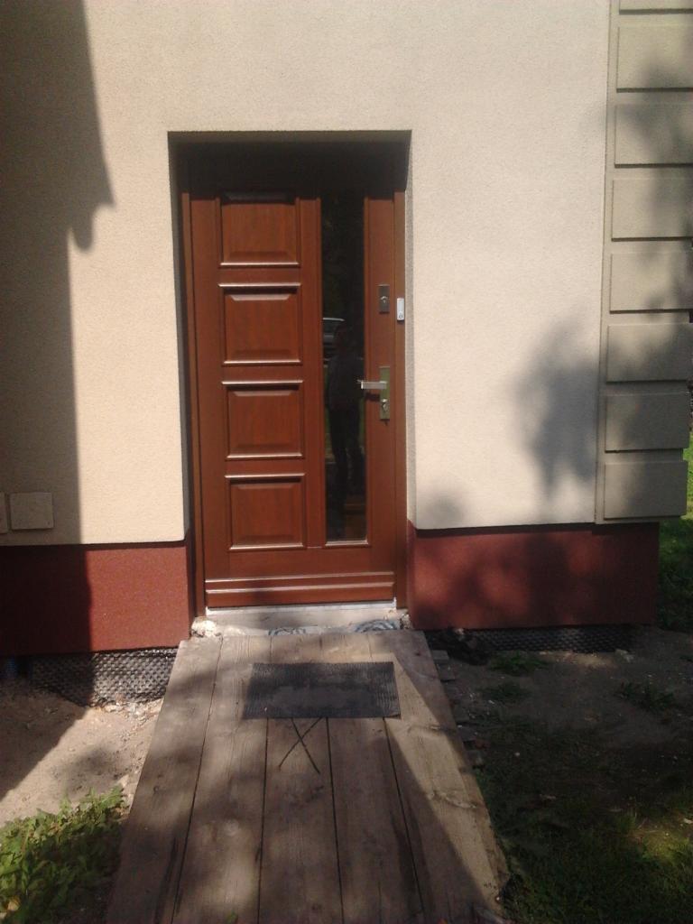 Częściowo otwarte, brązowe drzwi wejściowe z przeszkleniem i panelem, zamontowane w beżowej elewacji budynku z brązowym cokołem. Widoczne drewniane podesty.