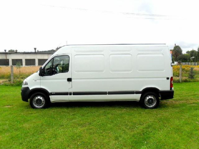 Biały bus dostawczy Opel Movano zaparkowany na trawie, widok z boku, z budynkiem w tle.