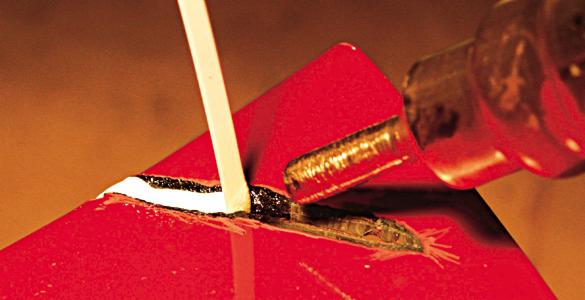 Detaliczne ujęcie procesu spawania plastiku na czerwonym elemencie z użyciem pręta spawalniczego i spawarki, widoczne ślady stopionego materiału.