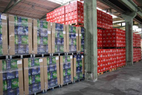 Palety z zapakowanymi skrzynkami piwa i czerwonymi skrzynkami ułożone w magazynie, widoczne betonowe kolumny.