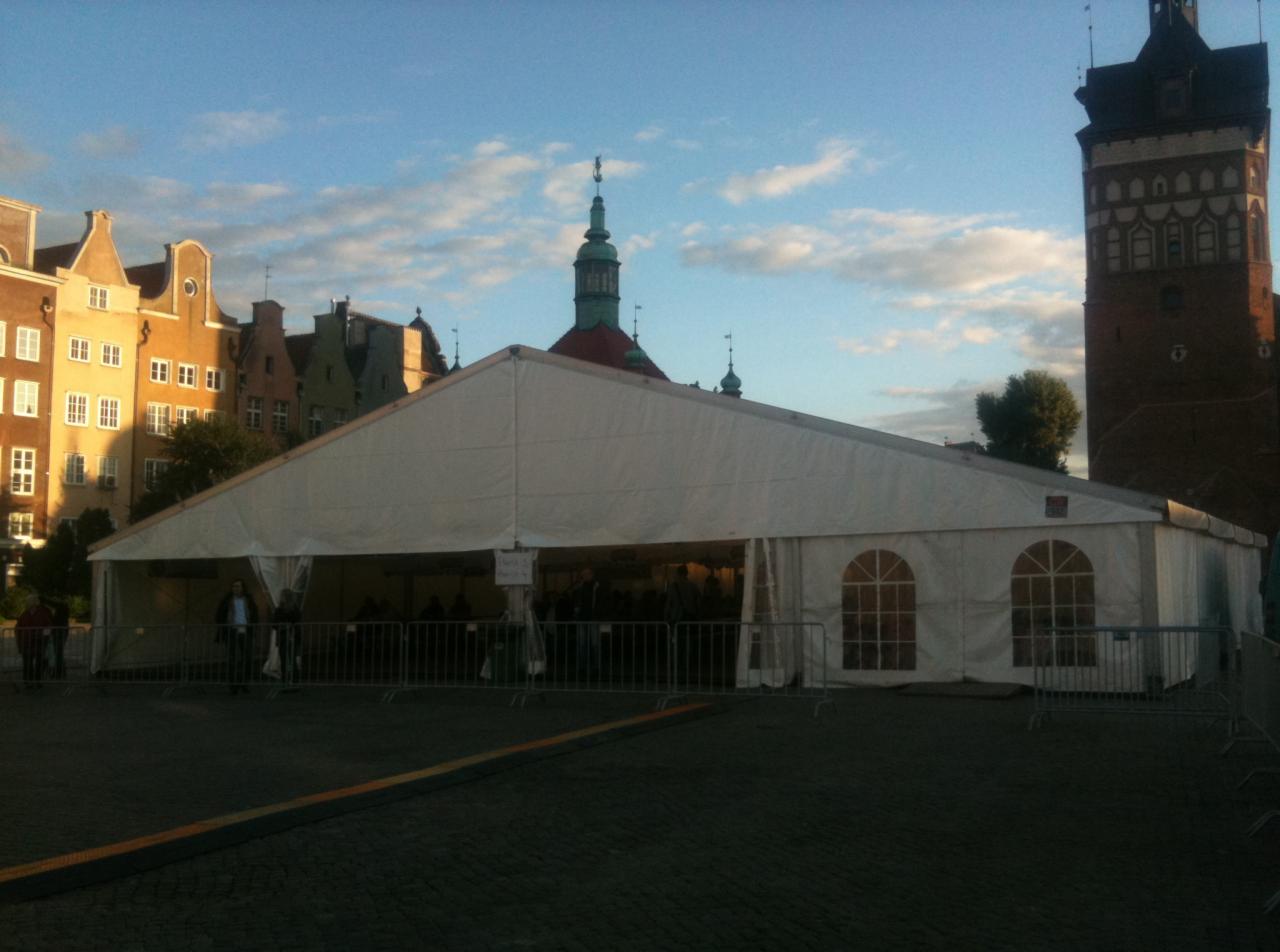Duży, biały namiot eventowy na tle historycznej zabudowy Gdańska, z widoczną wieżą ratusza i fragmentem Długiego Targu. Namiot zabezpieczony metalowymi barierkami.