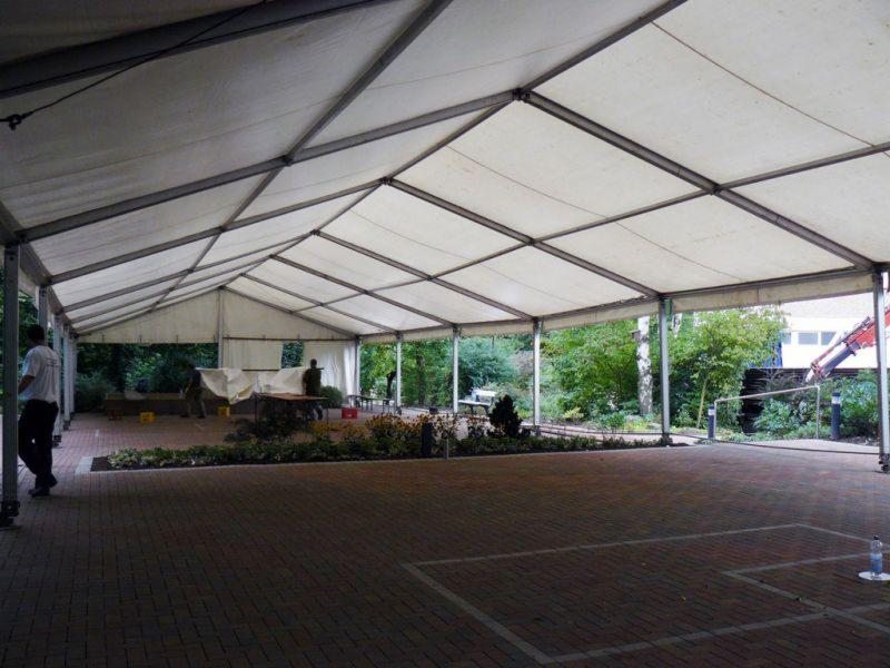 Wnętrze dużej hali namiotowej z widoczną konstrukcją stalową i białym poszyciem, częściowo otwartej na zewnątrz, z widocznymi osobami przygotowującymi przestrzeń i fragmentem krajobrazu z zielenią...
