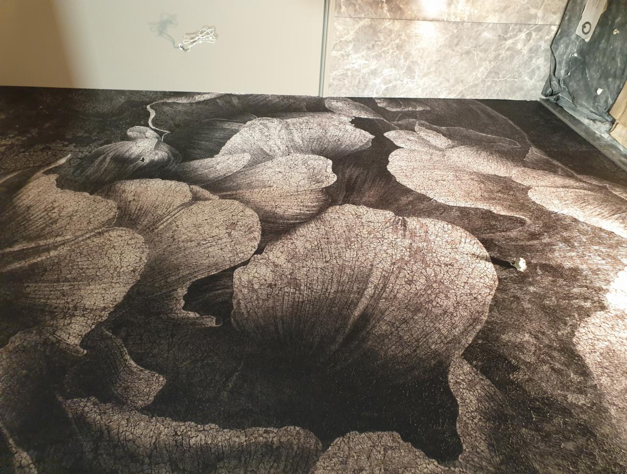 Podłoga po położeniu tapety z motywem kwiatowym w odcieniach szarości, widoczne łączenia i fragment ściany z wystającymi przewodami.