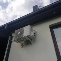 Zewnętrzna jednostka klimatyzacji marki Kaisai zamontowana na ścianie budynku z widocznym oknem i orynnowaniem, na tle pochmurnego nieba.