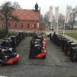 Rząd nowych wózków widłowych Linde i Fenwick na brukowanym placu, z budynkiem z czerwonej cegły i flagami Profit Partner w tle, pochmurny dzień.