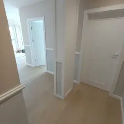 Jasny korytarz po remoncie z drewnianą podłogą, białą stolarką drzwiową i dekoracyjnymi panelami ściennymi w odcieniach bieli i szarości.