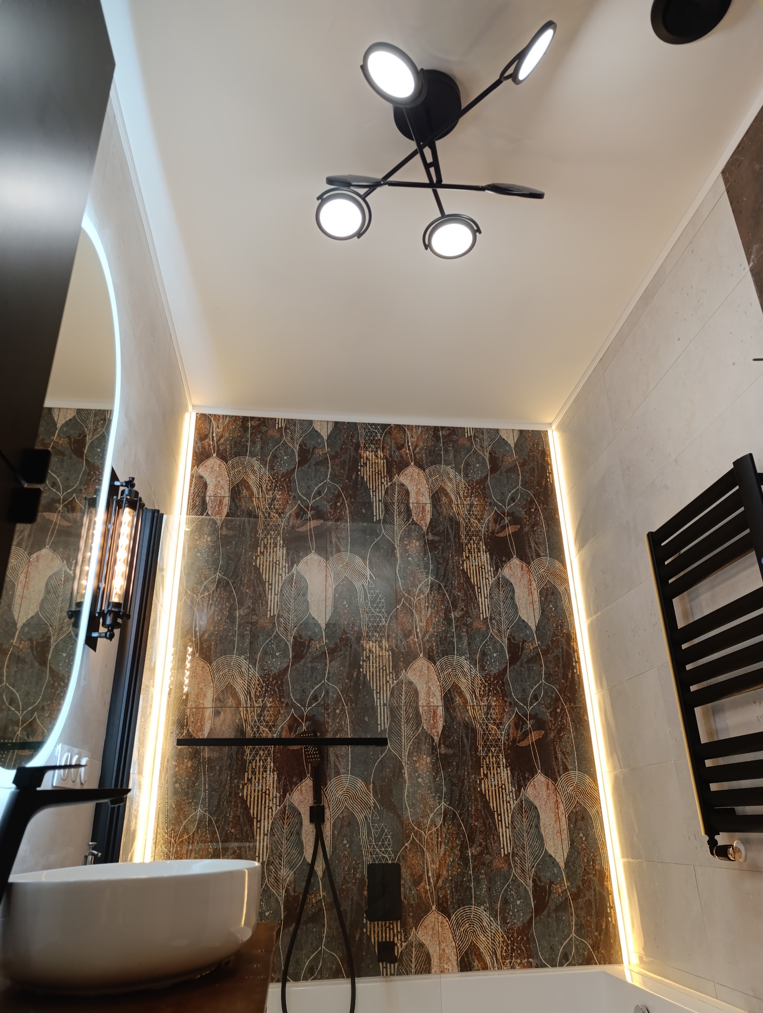 Nowoczesna łazienka z umywalką nablatową, baterią w kolorze czarnym, dekoracyjnymi płytkami na ścianie, podświetleniem LED i designerską lampą sufitową.