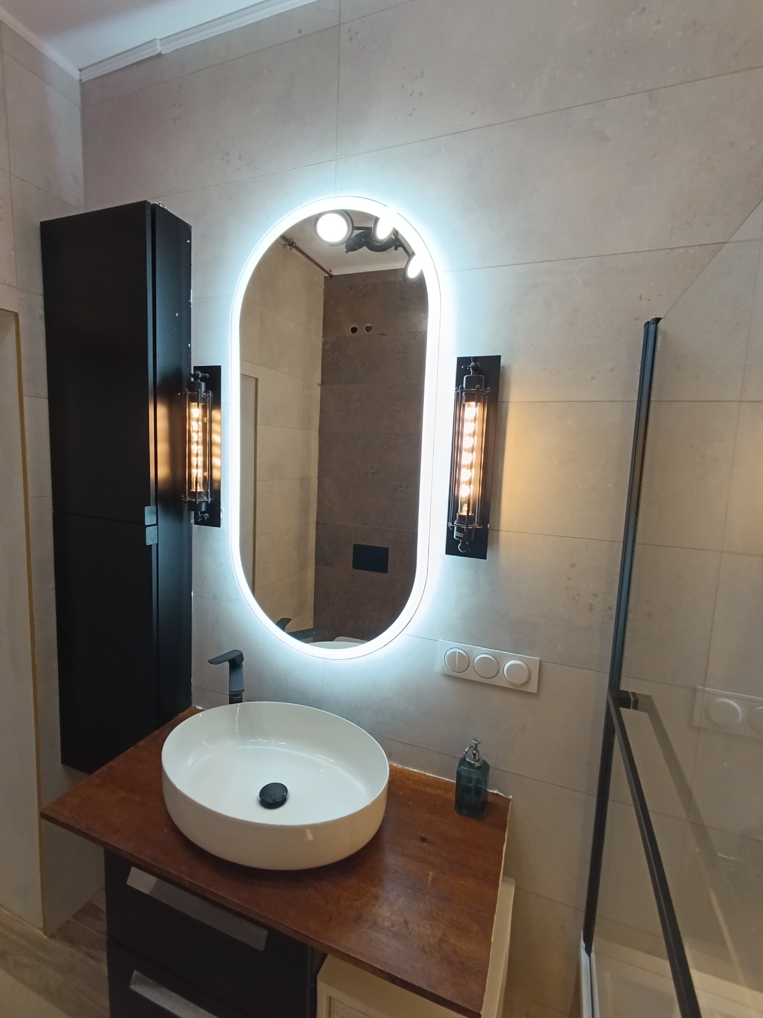 Nowoczesna łazienka z owalnym lustrem podświetlonym LED, umywalką nablatową na blacie z ciemnego drewna i industrialnymi kinkietami po bokach lustra.