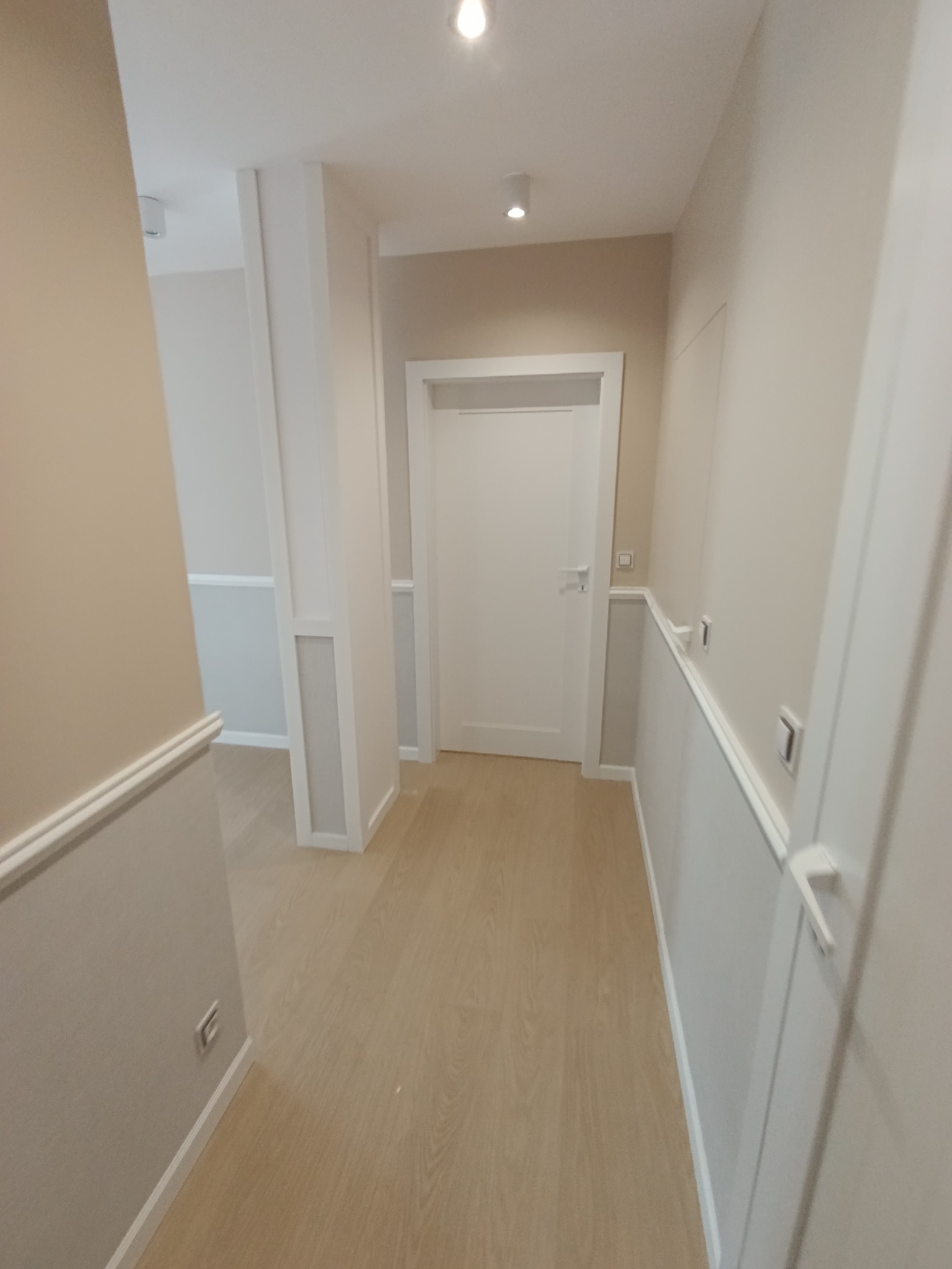 Korytarz po remoncie w neutralnych barwach, z drewnianą podłogą, listwami przypodłogowymi i sztukaterią ścienną, zakończony białą, zamkniętą ścianą.