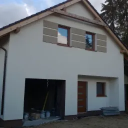 Dwukondygnacyjny dom w trakcie wykańczania elewacji, widoczne puszki z farbą, drewniane elementy konstrukcji dachu i dekoracyjne panele elewacyjne wokół okien.