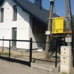 Nowoczesny dom jednorodzinny z białymi ścianami i czarnym dachem, widoczny zza czarnego, metalowego ogrodzenia panelowego z betonową podmurówką, z żółtą tablicą informacyjną na słupie w tle.