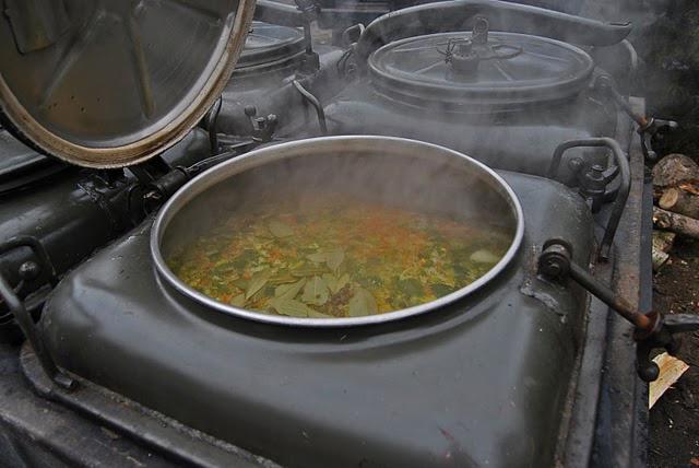 Kocioł polowy z otwartą pokrywą, wewnątrz gotująca się zupa z liśćmi laurowymi, para unosi się w powietrzu, w tle inne kotły i drewno.