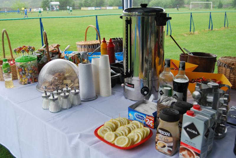 Stół z białym obrusem, na którym ustawiono automat do kawy, naczynia, przekąski i napoje; w tle boisko sportowe z grającymi osobami.