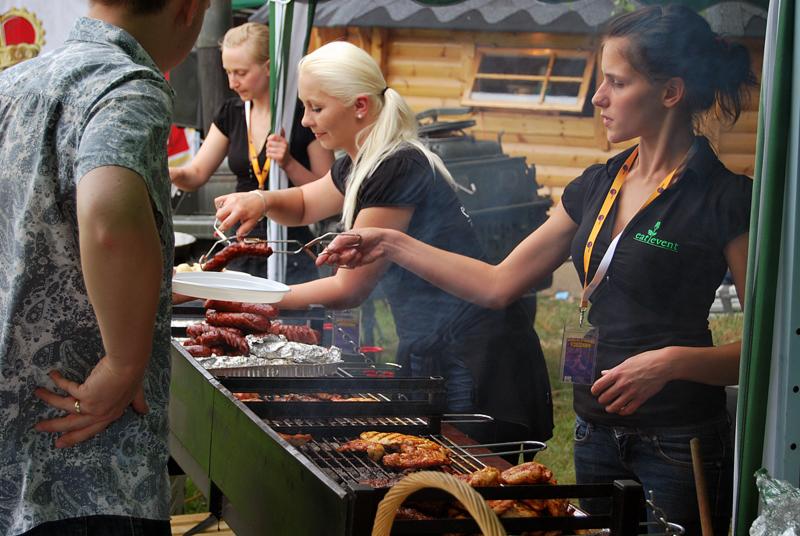 Obsługa cateringu podczas imprezy plenerowej: trzy kobiety w czarnych koszulkach serwują grillowane kiełbaski i mięso z rusztu na talerze dla gości. W tle drewniana altana.