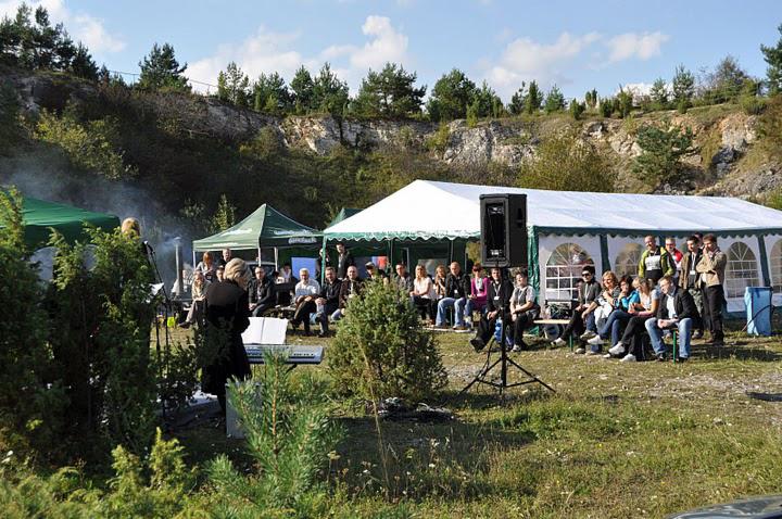 Plenerowa impreza firmowa z białym namiotem bankietowym, sceną muzyczną i publicznością siedzącą na ławkach, w tle zbocze z zielenią.
