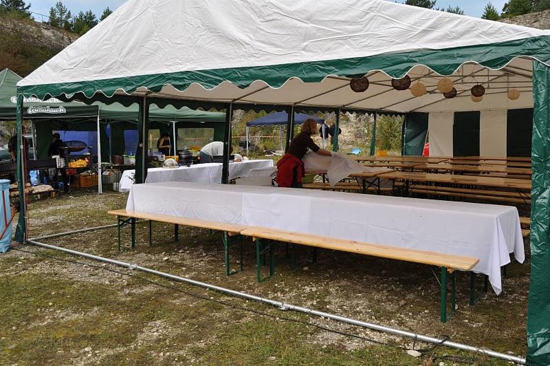 Przygotowania do imprezy plenerowej: rozkładanie białych obrusów na długich stołach pod namiotem bankietowym z zielonymi akcentami.