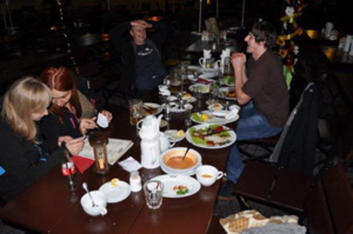 Grupa osób siedzi przy stole w restauracji po posiłku, widoczne resztki jedzenia na talerzach, puste naczynia i napoje, oświetlenie sztuczne, w tle dekoracja.