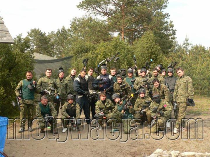 Grupa około 25 osób w strojach moro i maskach paintballowych, pozujących z markerami na poligonie leśnym, widoczny napis motywacja.com.
