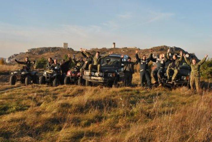 Grupa osób świętuje z podniesionymi rękami na tle dwóch samochodów terenowych i trzech quadów w plenerze z pagórkowatym krajobrazem w tle.