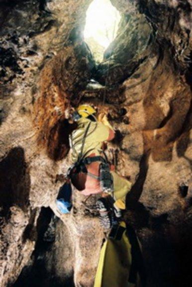Osoba w kasku i uprzęży zjeżdża na linie w dół jaskini, widoczne formacje skalne i otwór w stropie jaskini wpadające światło.