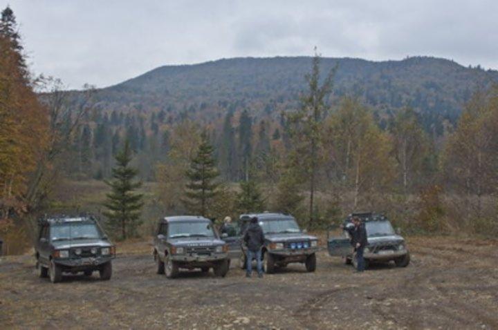 Cztery samochody terenowe Land Rover na tle zalesionych wzgórz podczas Bieszczady Trophy + Lwów, osoby w pobliżu pojazdów.