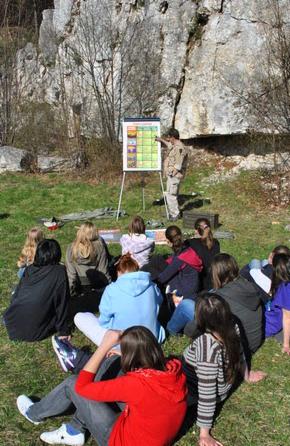 Grupa młodzieży siedząca na trawie słucha wykładu instruktora przed tablicą poglądową na tle skalistego krajobrazu Jury Krakowsko-Częstochowskiej.
