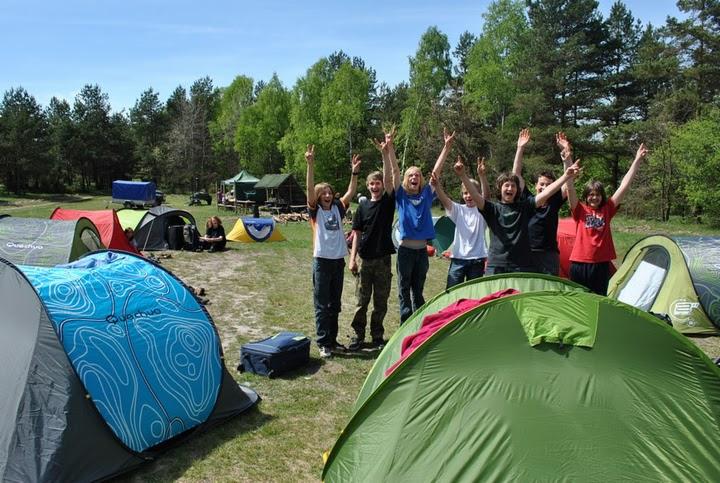 Grupa dzieci z podniesionymi rękami pozuje na tle rozbitego obozowiska z kolorowymi namiotami podczas zielonej szkoły na Jurze Krakowsko-Częstochowskiej.