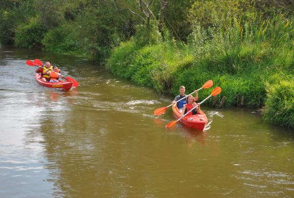 Dwa czerwone kajaki płynące pośród zielonej roślinności w brązowej rzece Warta, uczestnicy spływu kajakowego.