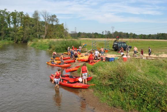 Grupa osób przygotowuje się do spływu kajakowego na rzece Warta, z czerwonymi i pomarańczowymi kajakami na brzegu, widoczne osoby w kamizelkach ratunkowych, na drugim planie łąka z zaparkowanym...