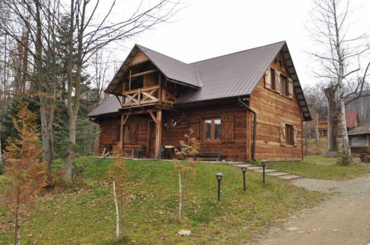 Rustykalny, drewniany dom z balkonem w Bieszczadach, z brązowym dachem i okiennicami, otoczony drzewami i trawnikiem z oświetleniem.