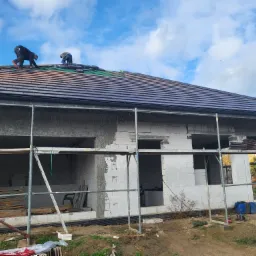 Dwóch dekarzy montuje dachówkę na nowym domu w stanie surowym, widoczne rusztowania i materiały budowlane.