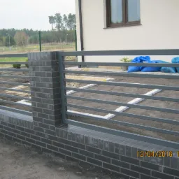 Nowoczesne ogrodzenie z szarej cegły klinkierowej i metalowych przęseł, widoczne niebieskie worki i przygotowany teren pod nawierzchnię.