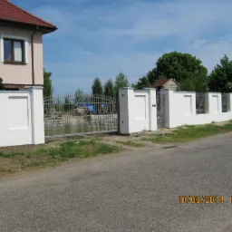 Nowoczesne ogrodzenie z białymi, prostokątnymi słupkami i metalową, ozdobną bramą wjazdową, widok z zewnątrz na tle domu i zieleni.