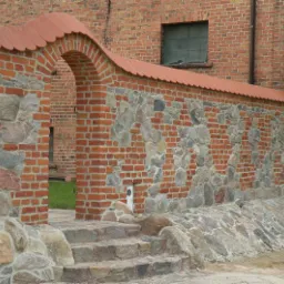 Mur z czerwonej cegły i kamienia polnego z łukowatym przejściem, wykończony czerwoną dachówką ceramiczną, prowadzący do zielonego trawnika, z widocznymi kamiennymi schodami i młodym drzewkiem.