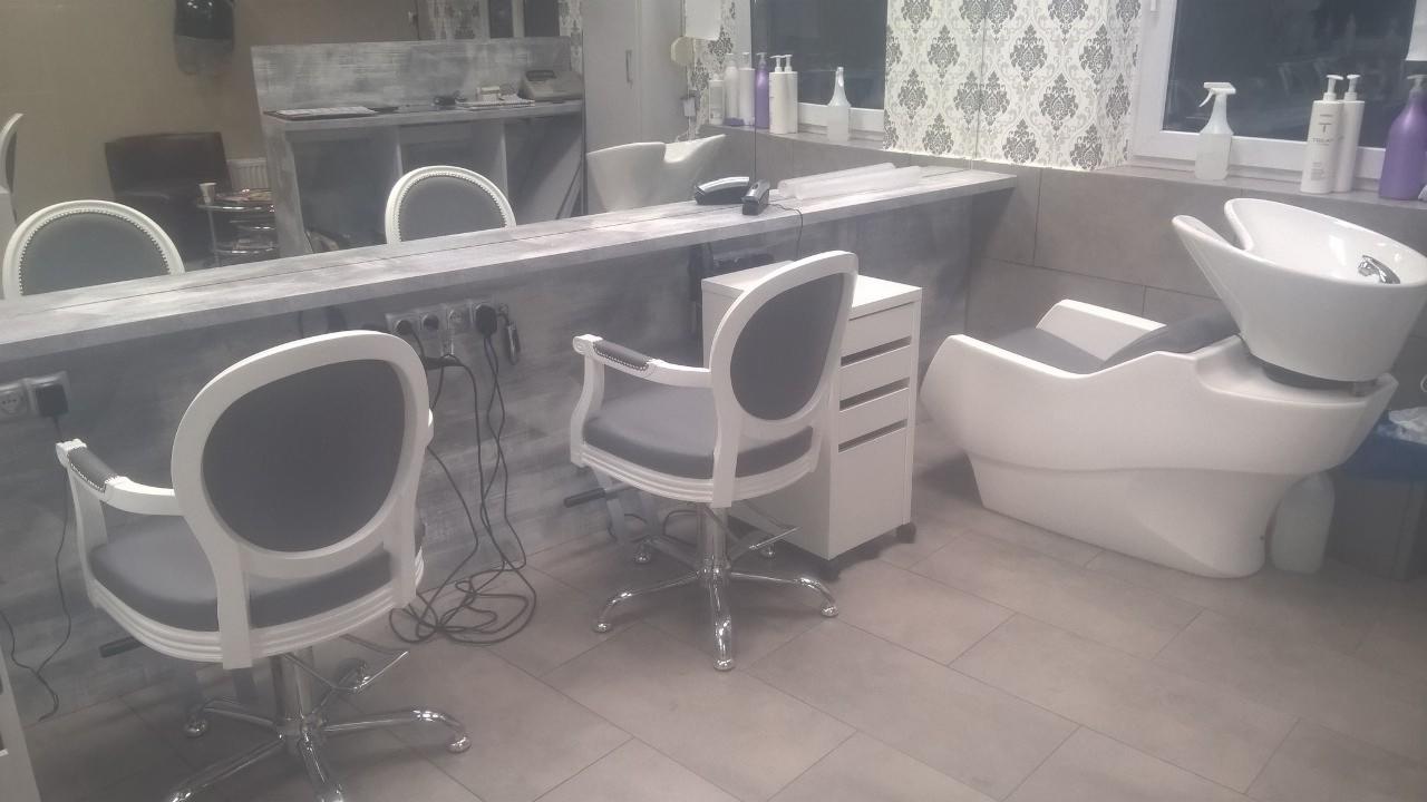 Wnętrze salonu fryzjerskiego z trzema białymi fotelami z szarymi siedziskami, białą myjnią fryzjerską i szarą ladą z lustrem, w tle tapeta w delikatny wzór.