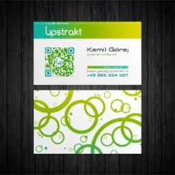 Wizytówka grafika komputerowego Kamila Góraja z logo Upstrakt, kodem QR i zielonym wzorem okręgów na białym tle, ułożona na ciemnym drewnianym podłożu.