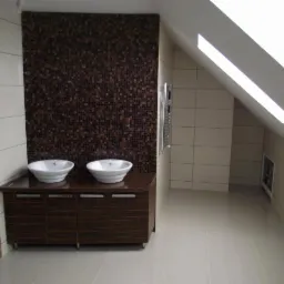 Nowoczesna łazienka na poddaszu z dwoma umywalkami nablatowymi, brązową mozaiką na ścianie i grzejnikiem drabinkowym, wykończona jasnymi płytkami ceramicznymi.