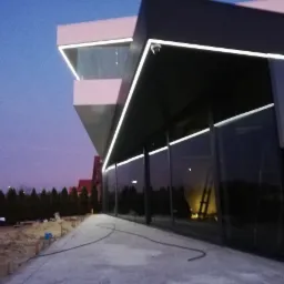 Nowoczesny budynek z elementami żelbetowymi i dużymi oknami, oświetlony listwami LED, widoczny na tle wieczornego nieba i drzew.