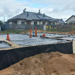 Budowa fundamentów domu jednorodzinnego: izolacja termiczna styropianem i folią, widoczne piony kanalizacyjne, w tle zabudowa mieszkalna i pracownik przy izolacji.