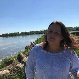 Portret kobiety w bluzce w paski na tle rzeki i zieleni w słoneczny dzień.