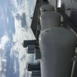 Zbiorniki gazowe LPG z żółtymi rurami na tle błękitnego nieba z chmurami. Widoczne metalowe konstrukcje i budynek w tle. Ujęcie z dołu, obrócone o 90 stopni.