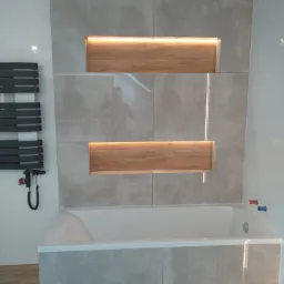 Nowoczesna łazienka z wanną obudowaną płytkami imitującymi beton, podświetlanymi półkami z drewnianym wykończeniem i grzejnikiem drabinkowym