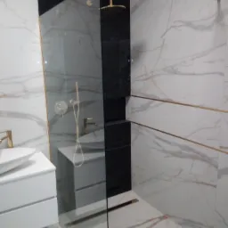 Nowoczesna łazienka z kabiną prysznicową typu walk-in, wykończona białym marmurem z delikatnymi brązowymi żyłkami, złotymi akcentami w postaci armatury i listew oraz czarnym panelem prysznicowym.