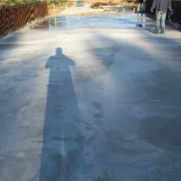 Świeżo wylana, gładka powierzchnia betonowa z widocznym zbrojeniem po bokach, cień osoby fotografującej w lewym dolnym rogu, dwóch pracowników w tle wygładza powierzchnię.