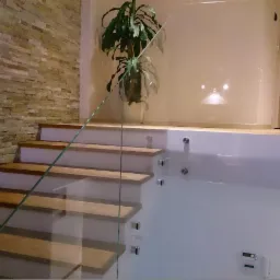 Nowoczesne schody z drewnianymi stopniami i szklaną balustradą, oświetlone ciepłym światłem, z dekoracyjną ścianą z kamienia w tle.