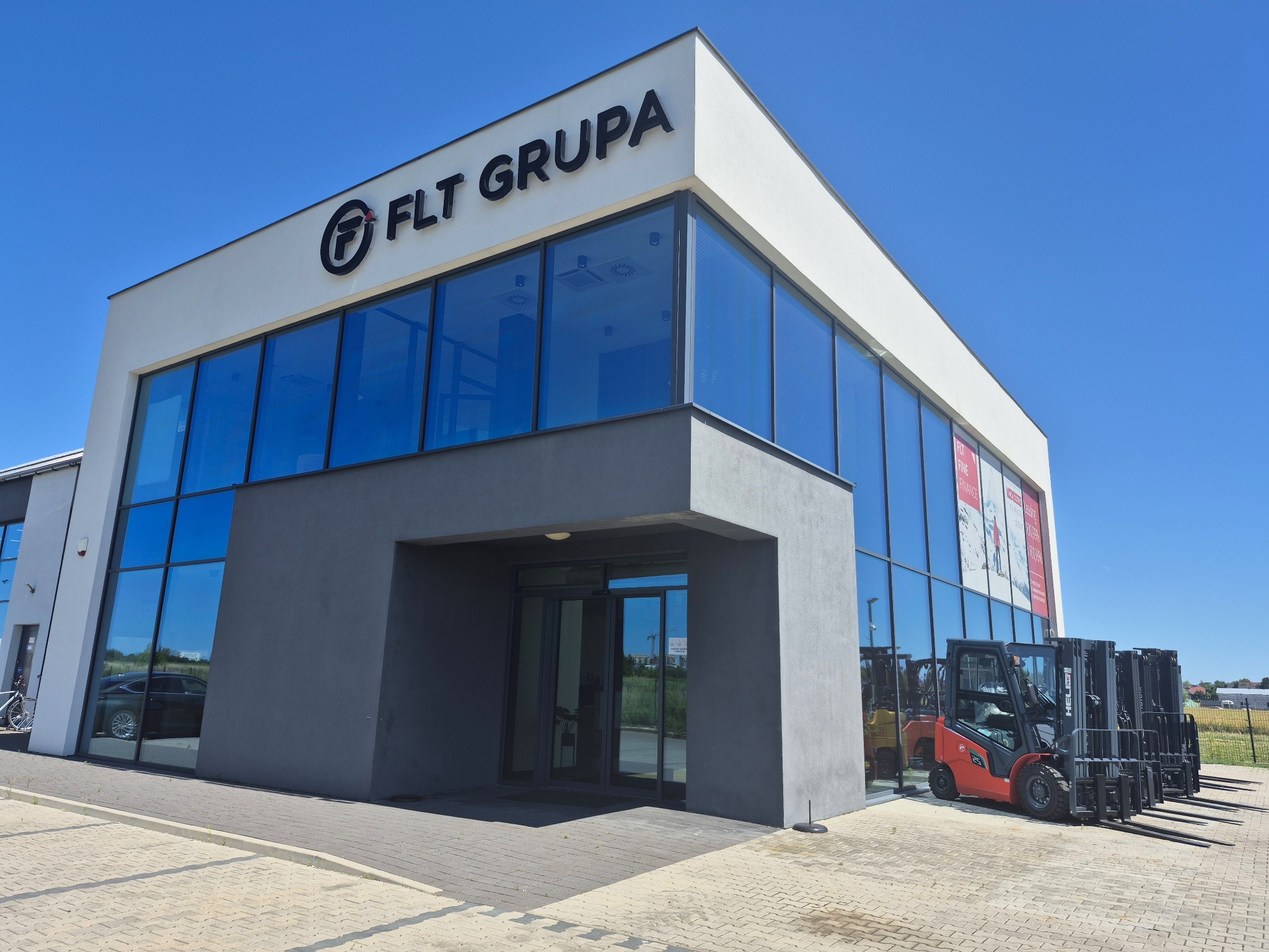 Nowoczesny budynek FLT GRUPA z przeszkloną fasadą, na zewnątrz ustawione wózki widłowe. Błękitne niebo w tle. Prezentacja firmy oferującej wózki widłowe.