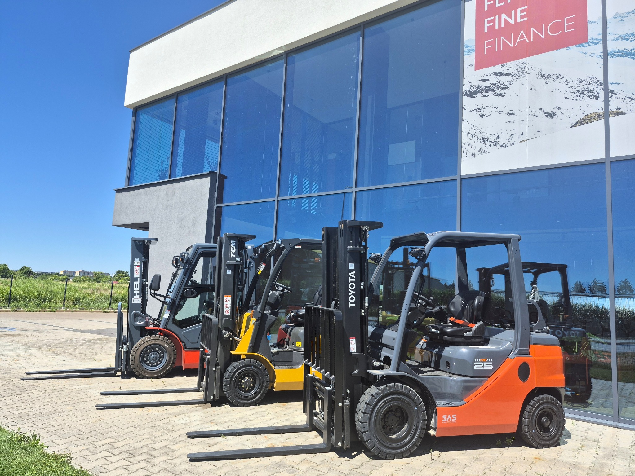 Trzy wózki widłowe (czerwony, żółty i pomarańczowy) stojące obok nowoczesnego budynku z dużymi oknami, w słoneczny dzień. Widoczny napis 'TOYOTA' na jednym z wózków.