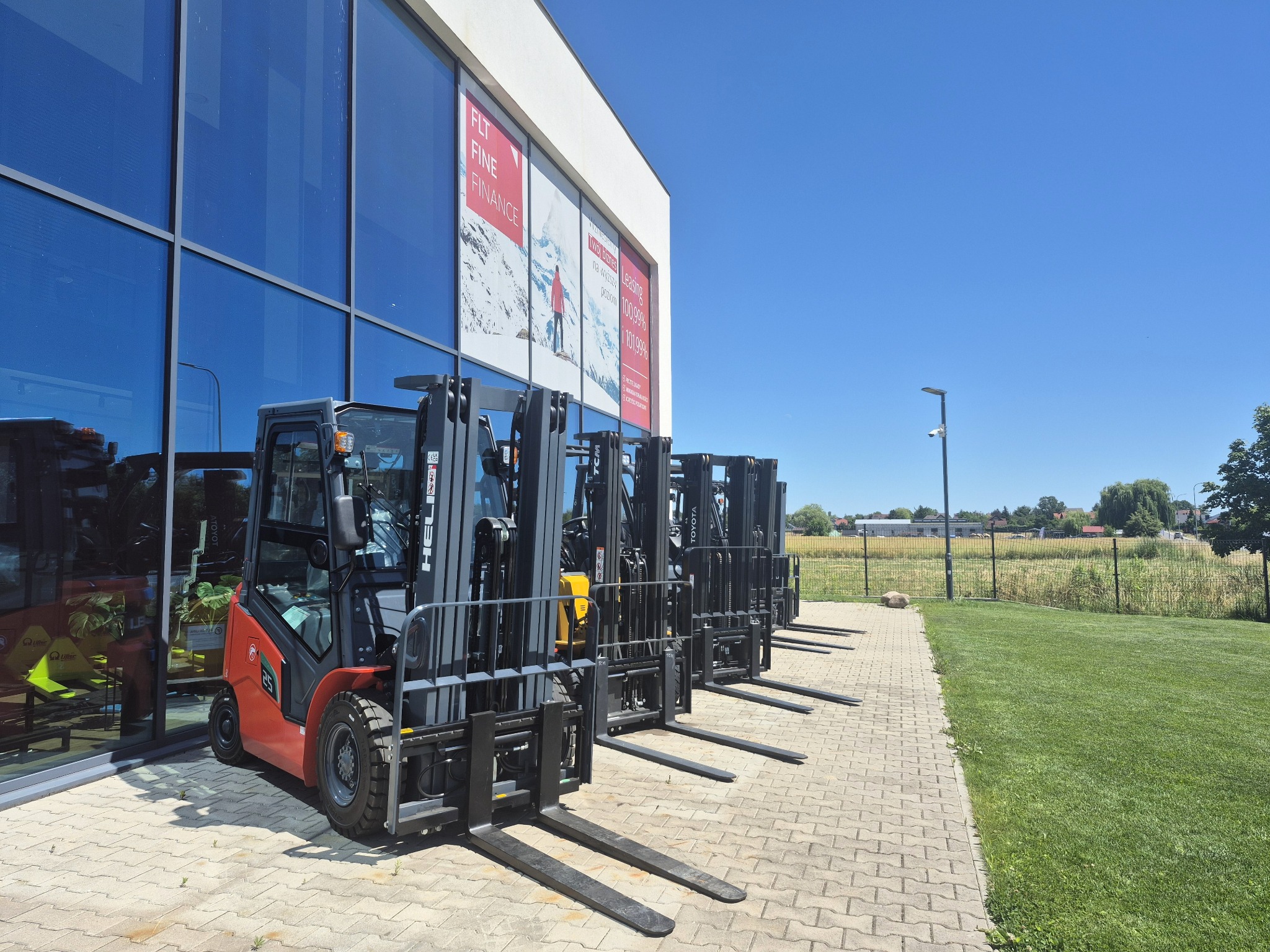 Rząd nowych wózków widłowych (czerwony HELI i czarne TOYOTA) przed budynkiem z dużymi oknami, w tle zielony trawnik i błękitne niebo.